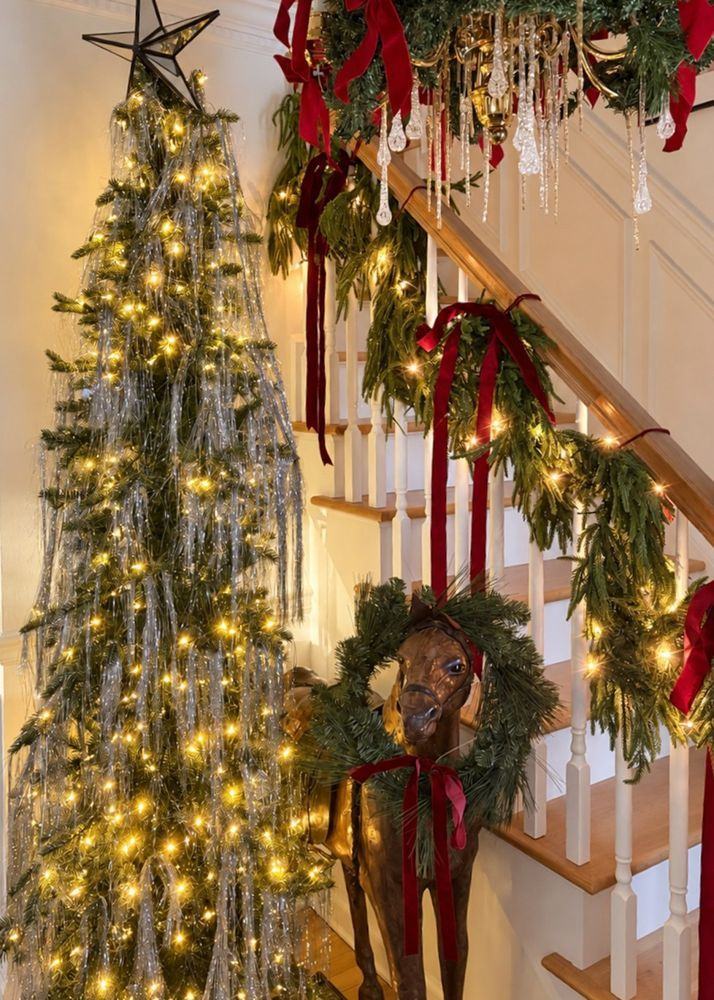 An entryway staircase wrapped in garland and red velvet ribbon beside a lit tree, with a wooden rocking horse wearing a wreath