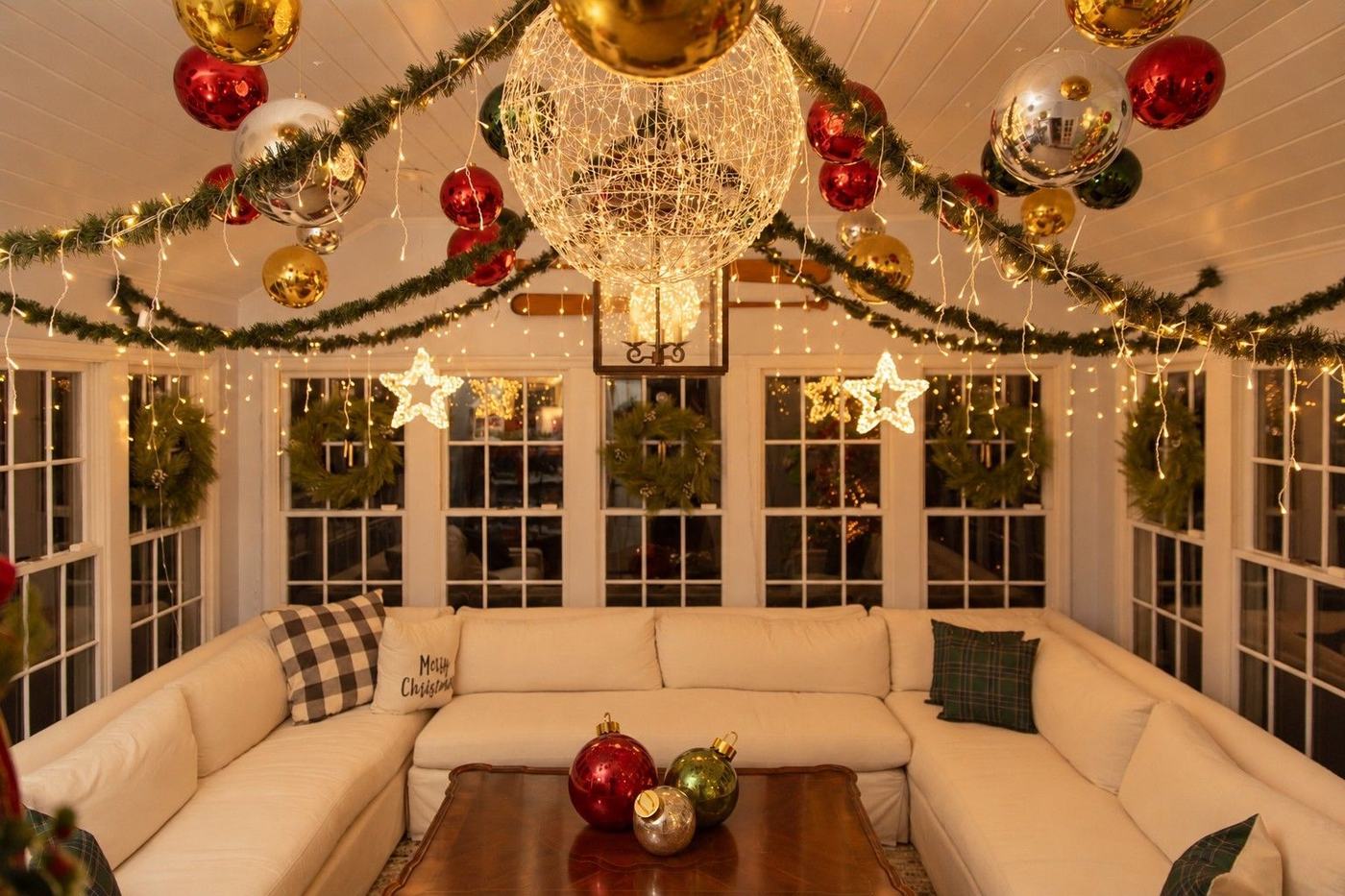 A garden room dressed for Christmas at dusk: ornaments and a glowing sphere overhead, wreaths in every window, a cream sectional