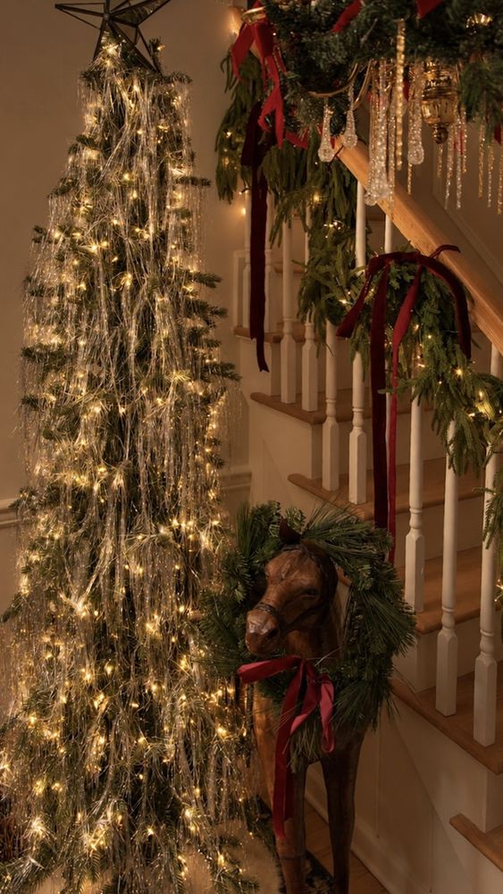A slim tree draped in silver strands and warm lights beside a garlanded staircase, a rocking horse wearing a wreath