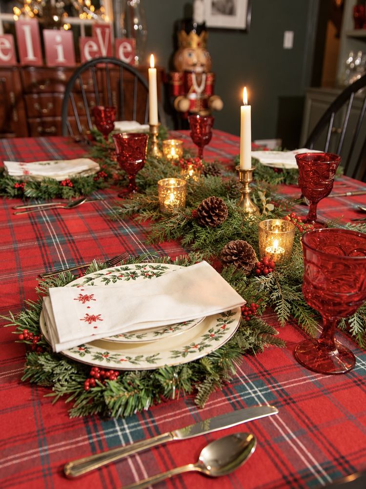 A tartan table set with holly china, red goblets, candles and a cedar runner, a nutcracker beyond
