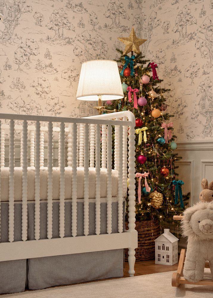 A nursery with toile wallpaper, a white spindle crib and a small lit tree hung with colorful ornaments and velvet bows