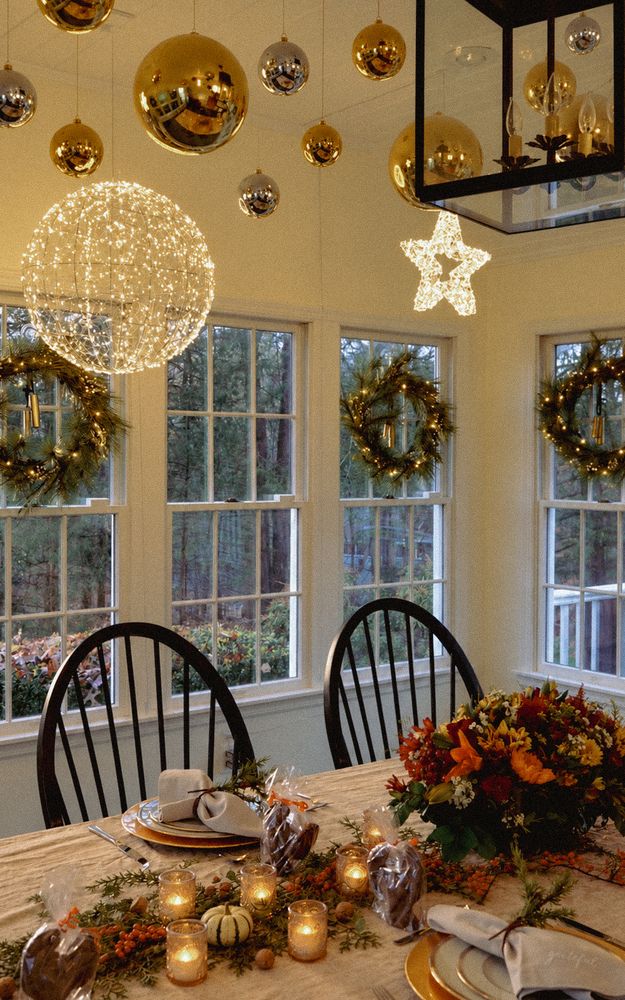 A Thanksgiving table in a sunroom beneath gold ornaments and a lit sphere, wreaths in every window