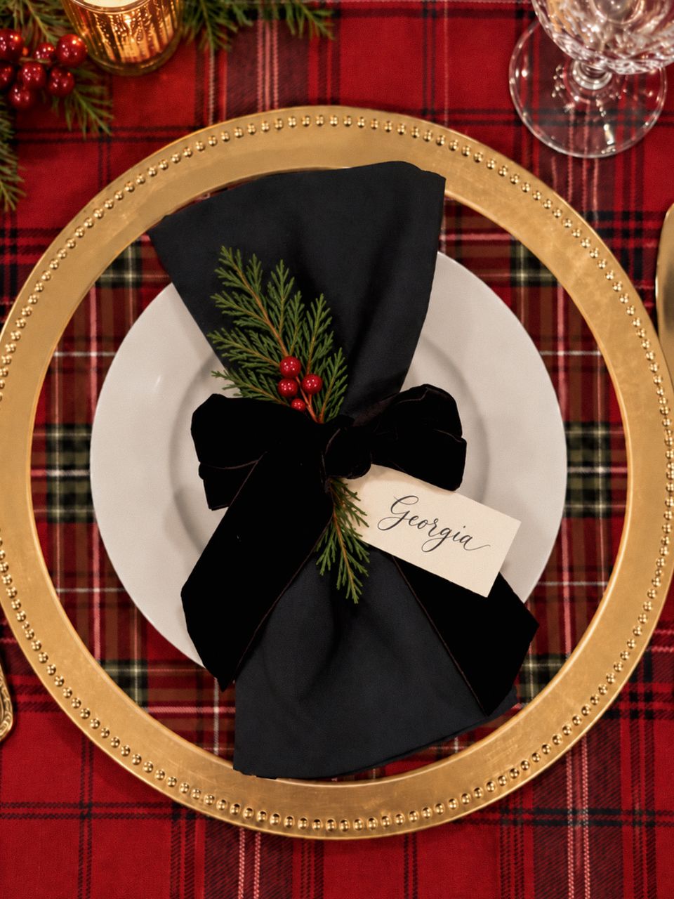 A place setting on a tartan tablecloth: gold beaded charger, black napkin tied with a velvet bow, cedar and berries, and a handwritten place card